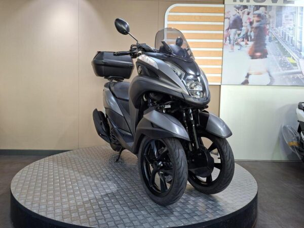 A sleek gray three-wheeled scooter displayed on a rotating platform in a showroom, with a backdrop featuring a blurred urban scene.