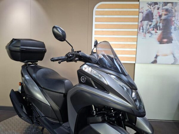 A stylish gray motorcycle with a black storage box mounted on the rear, set against a soft-focus background of people walking.
