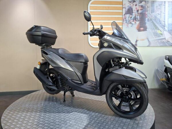 A sleek grey scooter displayed on a circular platform, featuring a top case, set against a minimalist indoor backdrop.