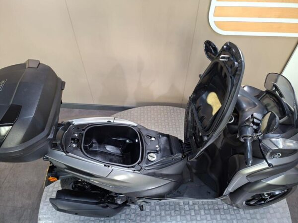 A grey scooter with an open seat revealing a storage compartment, parked on a textured floor against a plain background.