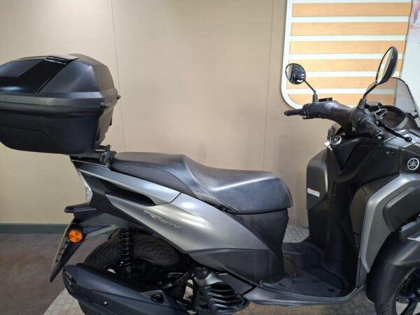 A motorcycle scooter with a sleek design and a black top case, parked on a textured floor in a neutral setting.