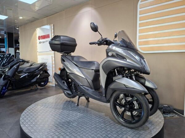 A Yamaha three-wheeled scooter displayed in a showroom on a circular platform, featuring a sleek gray design, a windscreen, and a rear storage box.