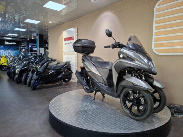 A gray three-wheeled scooter is prominently displayed on a circular platform in a showroom, surrounded by several black scooters in the background. The showroom features a Yamaha sign, with bright lighting and modern decor.