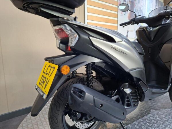 A close-up view of the rear of a black Yamaha Tricity scooter, showcasing the exhaust, suspension, and license plate (LC17 DRV), with a blurred background featuring a display of urban scenery.
