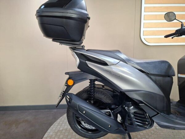 A side view of a silver scooter featuring a rear storage box, showcasing the suspension and rear wheel. The background is a neutral tone with a striped panel.