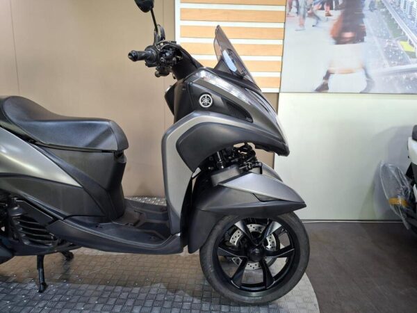 A gray three-wheeled scooter displayed in a showroom, featuring a sleek design, a clear windshield, and stylish black wheels. The background includes a light-colored wall with wooden accents.