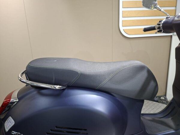 A close-up view of a scooter seat with a sleek design, featuring a dark fabric cover and chrome accents, set against a light-colored background.