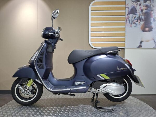 A blue Vespa scooter is displayed side-on in a modern showroom with soft lighting, featuring chrome mirrors, a comfortable black seat, and green accents on the body, set against a wall with a decorative wooden panel and a blurred photograph of pedestrians.