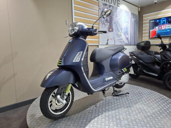 A sleek blue vintage scooter parked on a metallic display stand, with modern design elements and a stylish profile, set against a backdrop featuring a cityscape and a television screen.