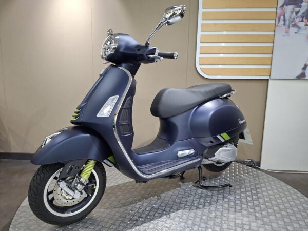 A sleek blue scooter with green accents is displayed on a textured surface, showcasing its modern design and chrome details against a neutral background.