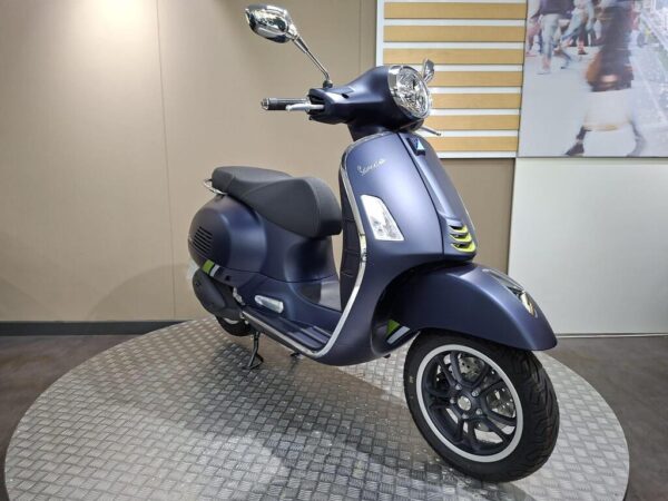 A blue matte Vespa scooter displayed on a circular metal platform against a beige wall backdrop. The scooter has a modern design with a distinctive front headlight and sleek lines.