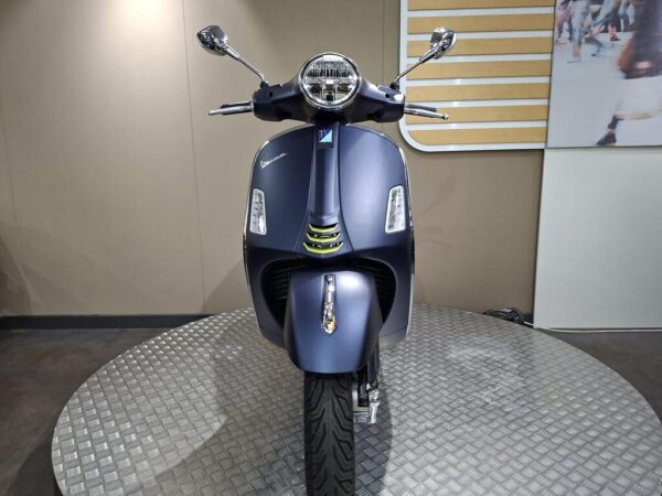 A front view of a blue Vespa scooter, featuring sleek lines, chrome accents, and a round headlight, displayed on a circular metal platform against a neutral background.