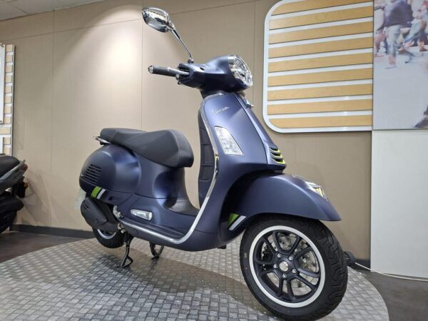 A blue Vespa scooter displayed in a showroom, featuring a sleek design with black seating and stylish alloy wheels, set against a modern, neutral-colored wall.