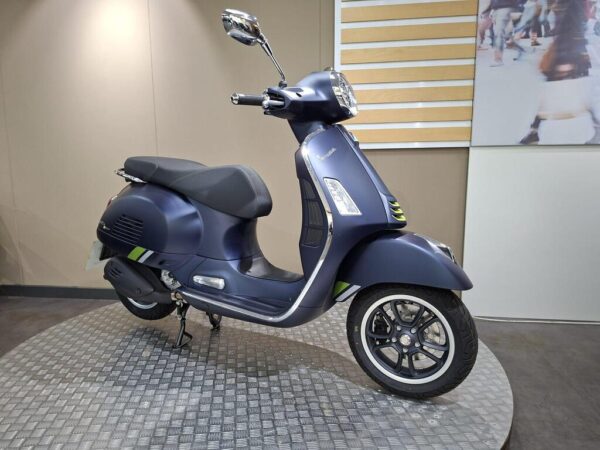 A blue Vespa scooter displayed in a showroom, featuring a sleek design with a black seat, alloy wheels, and subtle green stripes. The background includes neutral walls and a large image of a crowded street.