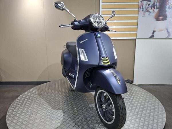 A dark blue Vespa scooter displayed on a rotating platform with a modern background, showcasing its sleek design and chrome accents.