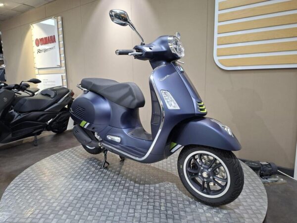 A blue Vespa scooter stands on a circular display, showcasing its sleek design and chrome accents, with a black motorcycle partially visible in the background.