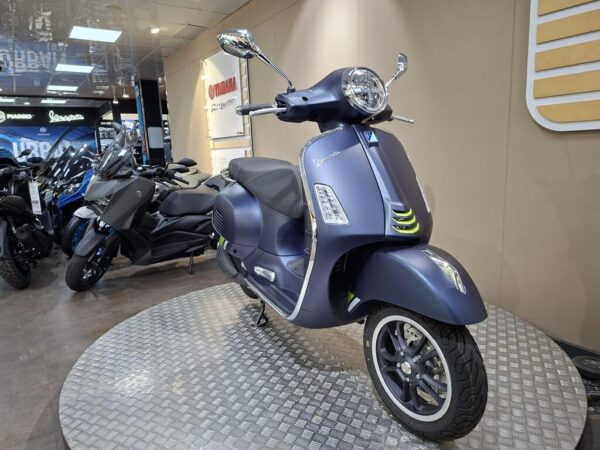 A stylish blue Vespa scooter displayed on a rotating platform in a motorcycle showroom, with various other bikes visible in the background.