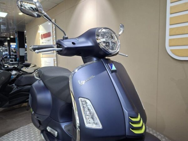 A sleek blue Vespa scooter is showcased in a modern display setting, featuring chrome mirrors, a circular headlight, and distinctive design elements such as an emblem and green accent near the front.