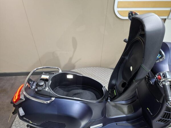 A close-up view of a scooter's storage compartment with the seat slightly lifted, showing a black, padded interior and controls on the side. The background features a neutral-colored wall.