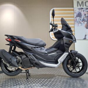 Aprilia SR GT 125 (2024) A black and silver scooter displayed on a textured circular platform, featuring a sleek design with a high front shield, a comfortable seat, and modern handlebars. The background includes a light-colored wall with a "MOVE" sign, and a blurred image of people walking.