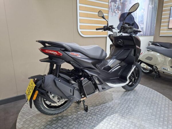 A black scooter displayed on a rotating platform, showcasing its sleek design and modern features, with a blurred background of a showroom highlighting other vehicles.