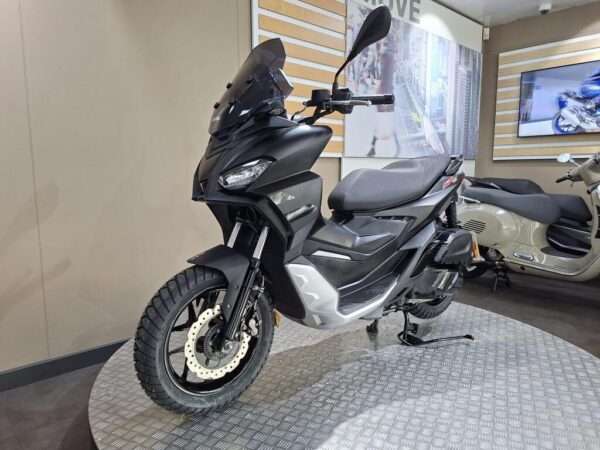 A black and silver scooter with a prominent windscreen is displayed on a textured platform in a modern showroom, with a light-colored scooter visible in the background.