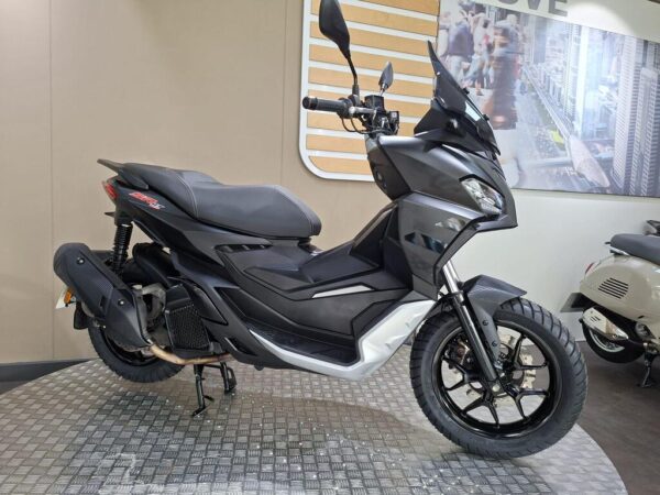 A sleek, modern black scooter displayed in a showroom, featuring a sporty design, front windshield, and textured seating. The background includes a cityscape image and other scooters.