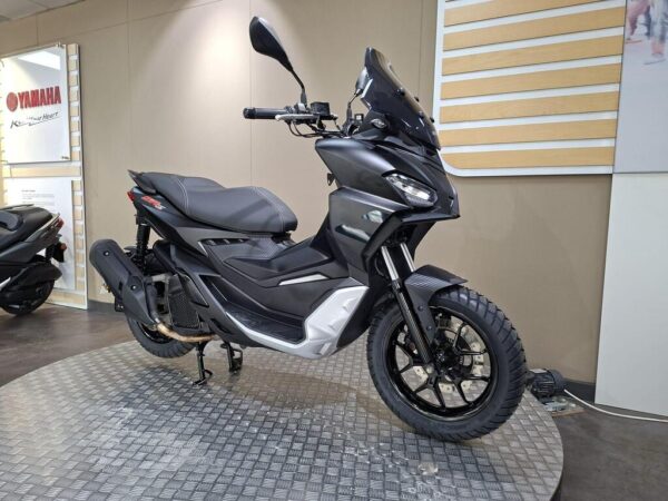 A sleek, black Yamaha scooter displayed in a showroom, featuring a modern design, off-road tires, and a stylish windscreen, with a light-colored patterned floor and minimalistic wall decor in the background.