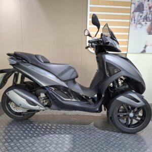A sleek grey three-wheeled scooter displayed on a circular platform with a simple background.
