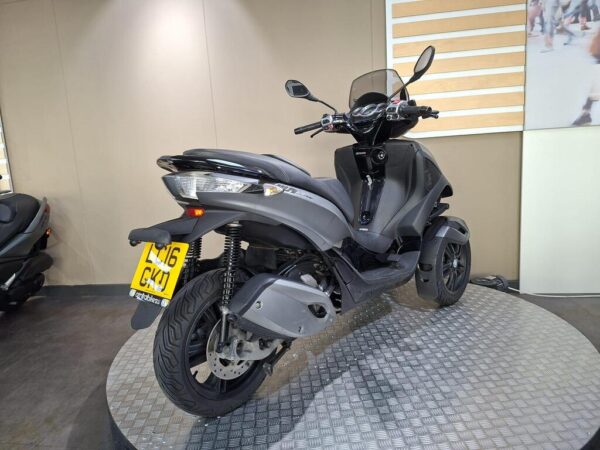 A gray three-wheeled scooter displayed on a circular platform in a showroom, featuring a modern design, front windshield, and visible license plate.