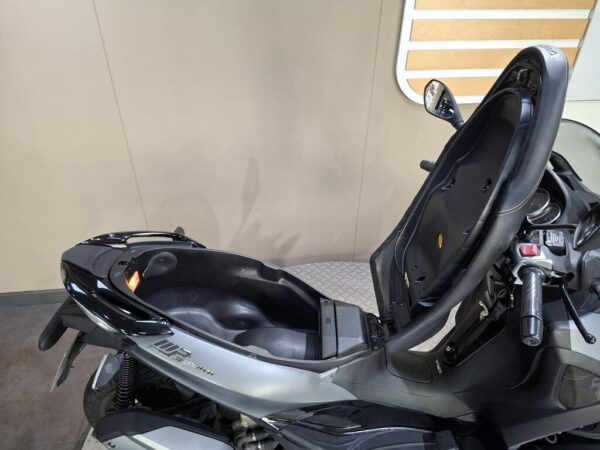 A close-up view of a black and silver scooter with the seat lifted, revealing storage space underneath, set against a neutral background.