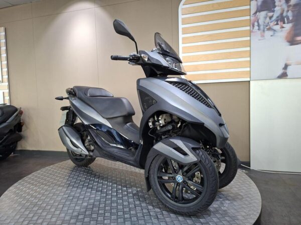 A sleek, gray three-wheeled scooter displayed on a circular platform in a modern showroom, featuring a streamlined design and a black seat. Soft lighting highlights its contours against a simple background.