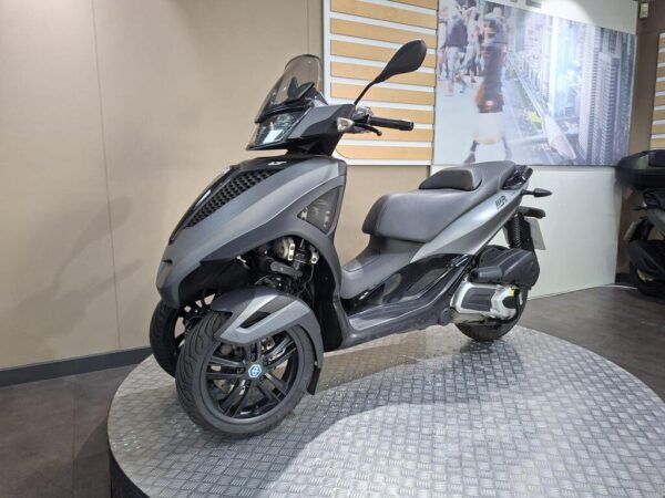 A sleek, modern three-wheeled scooter displayed on a circular platform, featuring a matte black finish, a streamlined design, and prominent front headlights. The background includes a blurred image of people walking in an urban setting.