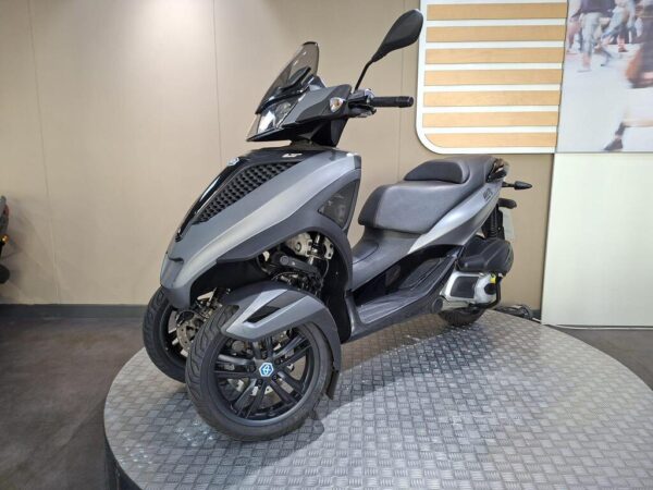 A sleek, modern three-wheeled scooter stands on a circular display pedestal, showcasing its gray body, sporty design, and large front windscreen. The background features a simple, light-colored wall with horizontal wooden slats.