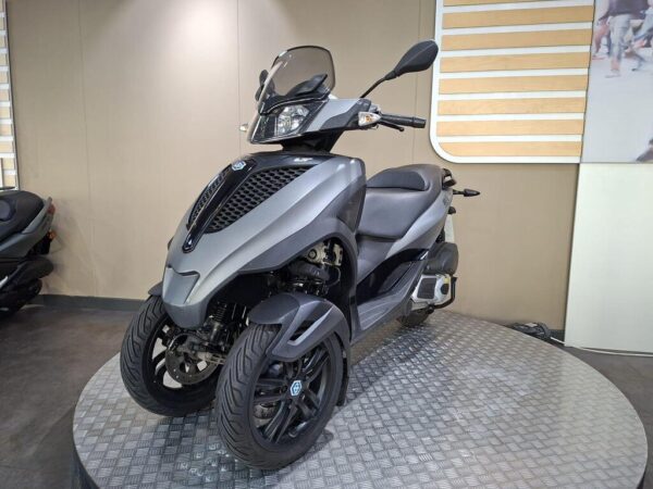 A sleek, modern three-wheeled scooter in metallic gray, parked on a circular platform with a textured surface. The scooter features a windscreen, stylish body design, and two front wheels with prominent tires. The background is neutral, emphasizing the scooter's design.