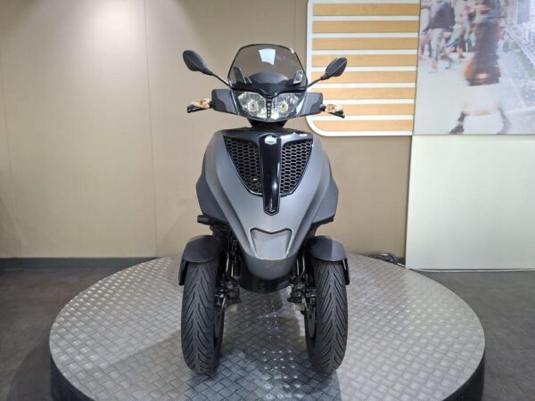 A gray three-wheeled scooter is displayed on a circular platform, facing forward, with its headlights on and a blurred background featuring a bustling urban scene.