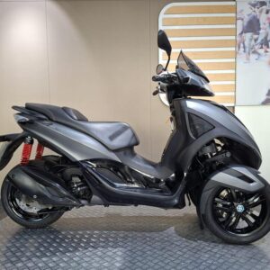 Piaggio MP3 300 (2012) A sleek, gray three-wheeled scooter displayed on a textured platform, featuring a comfortable seat, sporty design elements, and prominent rear suspension in red. The background includes a stylized wooden panel and a blurred image of people walking.