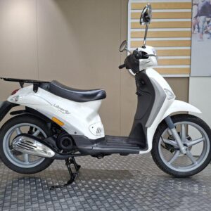 Piaggio Liberty 50 Delivery (2015) A white scooter is positioned on a patterned surface, displaying its side profile. The scooter features a black seat, chrome exhaust, and a rear luggage rack, with a neutral background that includes light-colored walls.
