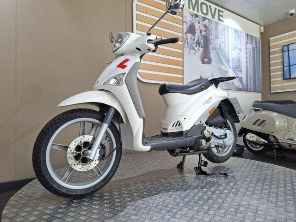 A white scooter with an "L" learner plate displayed on a checkered floor, set against a backdrop of a cityscape in a bright showroom.