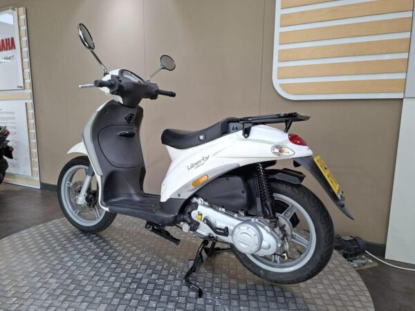 A white scooter parked on a textured floor, showcasing its sleek design, rear view, and prominent features like mirrors and a seat. The background includes a minimalist wall with wooden accents.