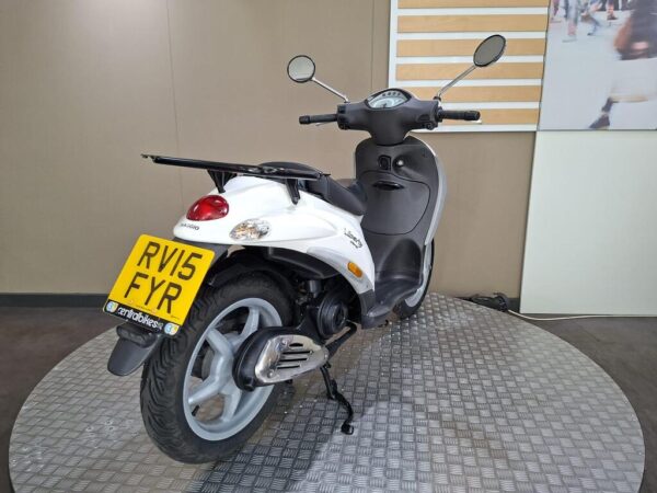 A white Piaggio scooter is displayed on a circular stand, featuring a rear view with a visible license plate reading "RV15 FYR." The background includes a neutral-colored wall and a partial view of an abstract image.