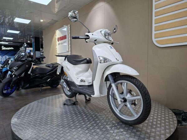 A white scooter is displayed on a circular platform in a showroom with a wooden and beige backdrop, alongside a black motorcycle.