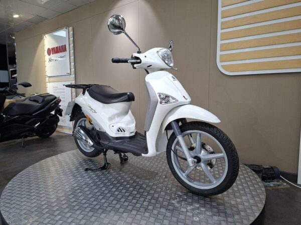 A white scooter displayed on a circular metal platform inside a Yamaha dealership, with a black scooter partially visible in the background.