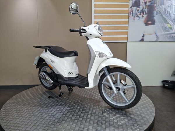 A white scooter displayed on a circular, metallic platform against a neutral background, featuring a sleek design with a single headlight and a comfortable seat.