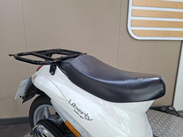 A close-up view of the rear section of a white scooter featuring a black seat and a metal luggage rack on top, set against a neutral-colored background.