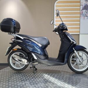Piaggio Liberty 50 (2019) A blue scooter with a black top case is displayed on a circular textured base, set against a neutral background.