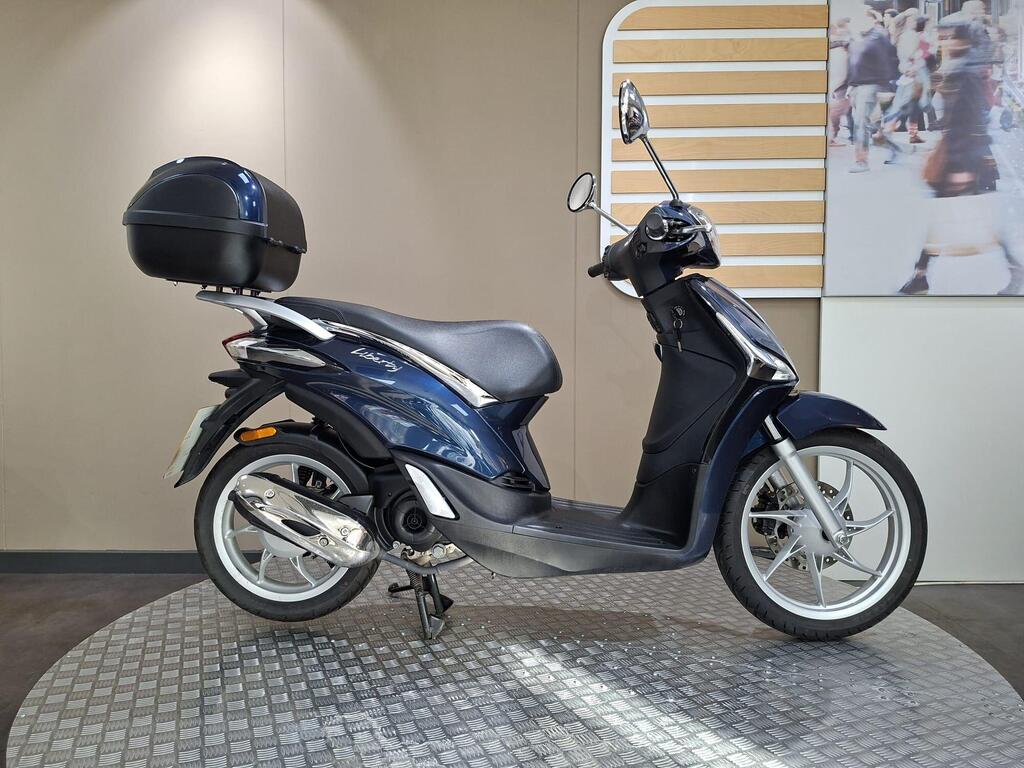 Piaggio Liberty 50 (2019) 1 Piaggio Liberty 50 (2019) A blue scooter with a black top case is displayed on a circular textured base, set against a neutral background.
