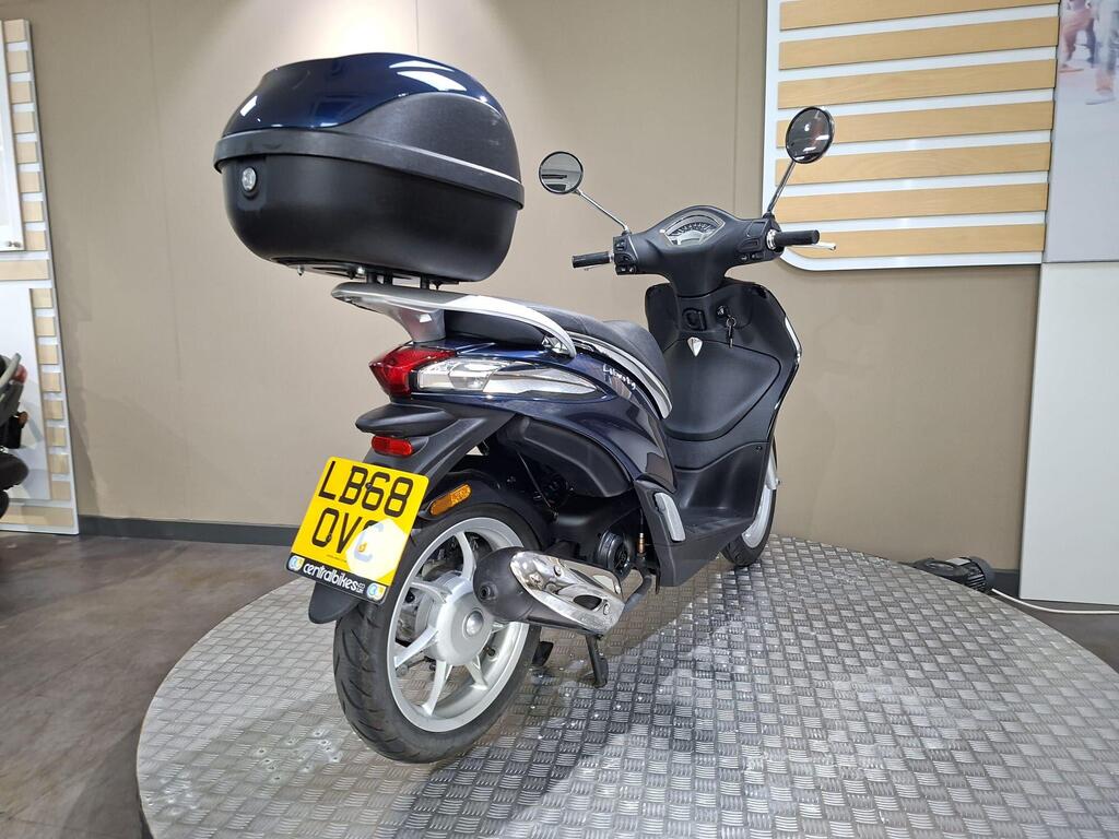 Piaggio Liberty 50 (2019) 11 A dark blue scooter with a top case stands on a display platform, showcasing its rear view. The license plate is visible, displaying "LB68 OQV." The background features a neutral-toned wall and a textured floor.