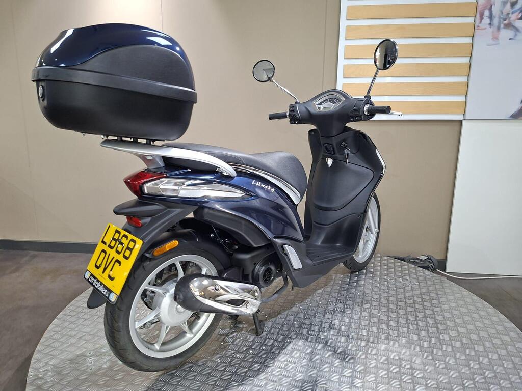Piaggio Liberty 50 (2019) 12 A dark blue scooter with a rear top case, parked on a textured circular platform, with a minimalistic indoor background.