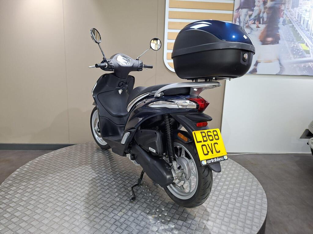 Piaggio Liberty 50 (2019) 13 A black scooter with a blue storage case on the rear, displayed on a circular platform, against a neutral background. The license plate is visible, reading "LB68 OVC."